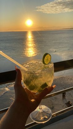 a hand holding a drink with a lime slice on the rim and water in the background