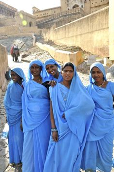 four women in blue dresses standing on the side of a road with an old building in the background