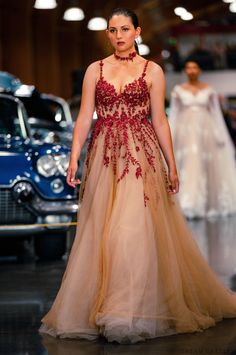 a woman is walking down the runway wearing a dress with red flowers on it and an old car in the background