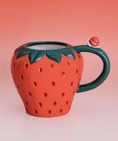 a ceramic strawberry shaped mug with a green handle on a pink background, holding a red berry