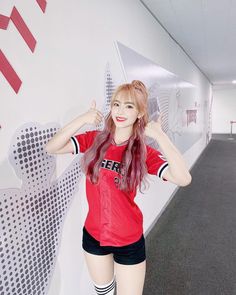 a woman with long pink hair standing in front of a wall and giving the thumbs up sign