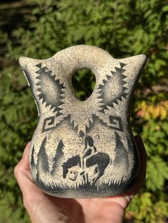 a hand holding up a black and white vase in front of some trees with green leaves