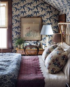 a bed sitting under a window next to a lamp and dresser in a room with blue wallpaper