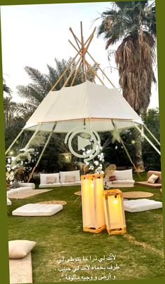 an outdoor tent set up in the grass with white pillows and lights on it, surrounded by palm trees