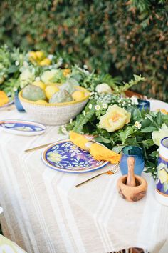 the table is set with plates, bowls and flowers on it for an outdoor meal