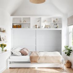 a white bed sitting under a window next to a shelf filled with books and vases