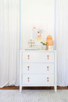 a white dresser sitting in front of a window next to a painting on the wall