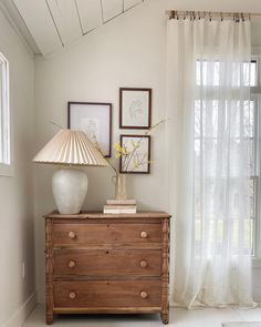 a white vase sitting on top of a wooden dresser next to a window with sheer curtains