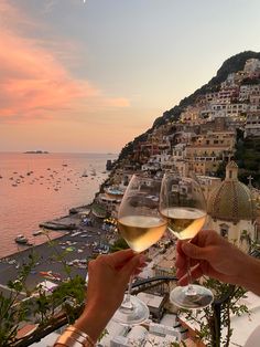 two people toasting with white wine on a balcony overlooking the ocean and town below