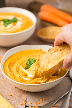 a person dipping bread into a bowl of soup with carrots and parsley on the side