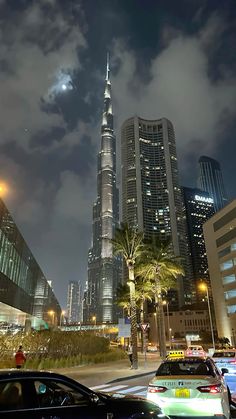 cars are parked on the street in front of tall buildings at night with lights and palm trees