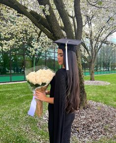 a woman with long hair wearing a graduation cap and gown holding flowers in front of a tree