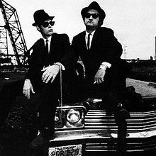 two men in suits sitting on the hood of a car with an oil rig in the background