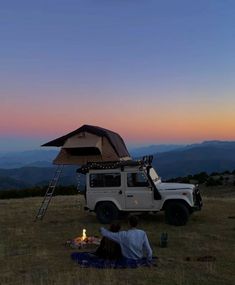 two people sitting in the grass under an awning next to a camper van