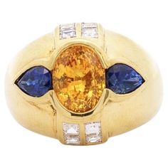 Vintage Yellow Sapphire with Blue Sapphire & Diamonds Dome Ring. This vintage dome ring is crafted from 18k yellow gold. The center stone is a 3 carat yellow sapphire with an oval-cut. Adorning the sides of the center stone are 0.75 carats of blue sapphire, two total. The ring also holds eight emerald-cut diamonds, weighing 1 carat total. All gemstones are secured with a bezel setting with an excellent symetry and color contrasting gemstones. Made with an evil eye motif. Item Details: - Type: Vi Yellow Gold Domed Sapphire Ring, Eye Motif, Dome Ring, Evil Eye Ring, Blue Sapphire Diamond, Yellow Gold Setting, Eye Ring, Domed Ring, Yellow And Blue