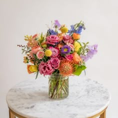 a glass vase filled with colorful flowers on top of a marble topped side table in front of a white wall