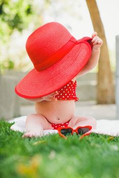 a baby wearing a red hat sitting on the grass with her hands in her hair