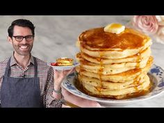 a man holding a plate full of pancakes with butter and syrup on it while standing in front of a stack of pancakes