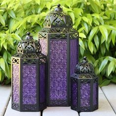 three purple lanterns sitting next to each other on a wooden table in front of bushes