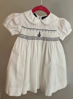 This blue and white Ralph Lauren dress is hand-smocked and the outer layer is made from delicately ribbed 100% white cotton. The smocking across the bodice features a darling anchor motif with white and blue striped  embroidery thread detailing. The voluminous sleeves elevate the overall look and have nice definition.  The garment is fastened using three (3) buttons up the back and a button belt that goes around the waist, (also fastened in the back). It's lined in feather-soft white cotton.  It White Fitted Smocked Dress For Baptism, Classic White Smocked Dress For Spring, Classic White Smocked Dress, Short Sleeve Smocked Dress For Baptism, White Lined Smocked Dress For Daywear, White Smocked Baptism Dress For Summer, White Smocked Dress For Baptism In Summer, White Smocked Dress For Summer Baptism, White Fitted Long-sleeve Smocked Dress