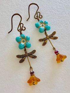 "Adorable turquoise blue Czech glass puffed daisies with topaz yellow bellflowers and vintage bronze stamped dragonfly charms. Matte turquoise 14 mm Czech glass puffy 5 petal flowers are etched with metallic gold and wire wrapped in vintage bronze with 6 x 9 mm translucent topaz yellow bellflowers accented with tiny burnt orange crystals. Lightweight dangles hang on handmade and hammered vintage bronze ear wires. Total drop length is 2 1/2\"." Whimsical Blue Jewelry With Flower Charm, Vintage Turquoise Dangle Flower Earrings, Blue Whimsical Dangle Flower Earrings, Whimsical Blue Dangle Flower Earrings, Nickel Free Turquoise Flower Earrings, Turquoise Nickel Free Flower Earrings, Handmade Turquoise Flower-shaped Earrings, Turquoise Dangle Earrings With Flower Charm, Vintage Blue Flower Earrings, Nickel-free