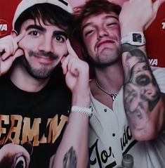 two men with tattoos posing for a photo in front of a red background, one is holding his hand up to the other's ear