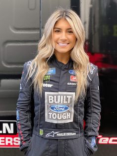 a woman with long blonde hair standing in front of a truck wearing a racing suit