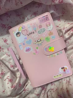 a pink notebook with stickers on it sitting on a bed next to a pen