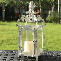 a white candle is sitting in a metal lantern on a table outside with grass behind it