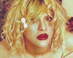 a woman with blonde hair and red lipstick wearing a flower in her hair is looking at the camera