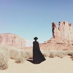 a person in a long black dress and hat standing in the desert with mountains in the background