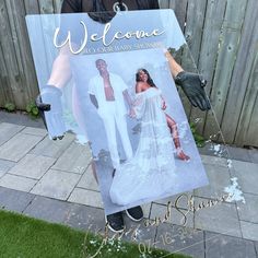 a man holding up a welcome sign in front of a wooden fence with grass and flowers