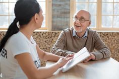 an older man sitting at a table talking to a young woman who is holding something in her hand