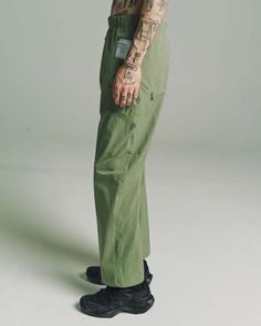 a tattooed man in green pants with tattoos on his arm and leg, standing against a gray background