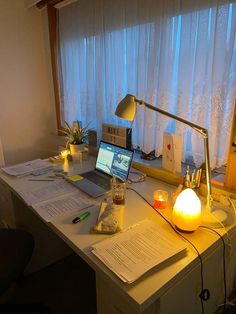 a laptop computer sitting on top of a white desk next to a lamp and papers