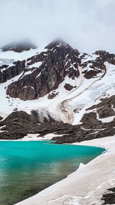 a snow covered mountain with a blue lake in the foreground and clouds above it