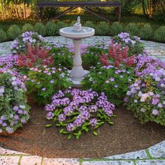 a fountain surrounded by purple flowers and greenery in the middle of a circular garden