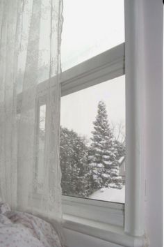 a bedroom window with white curtains and snow covered trees outside the window sill in front of it