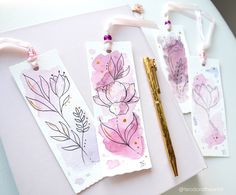 three watercolor bookmarks with pink flowers on them and a gold pen next to it