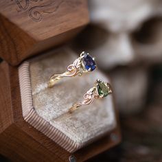 two gold rings with blue and green stones on them sitting in a wooden ring box