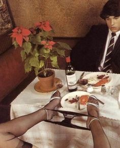 two people sitting at a table with food and wine in front of them, one person has their feet up on the table