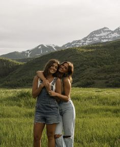 two women hugging each other in the middle of a field with mountains in the background