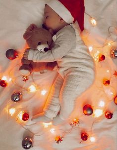 a baby in a santa hat is sleeping with a teddy bear and christmas lights on the bed