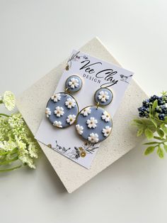 a pair of blue and white flower earrings on top of a card next to flowers