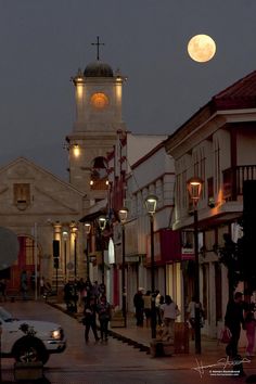 the full moon is setting over an old town with people walking on the sidewalk and cars driving down the street