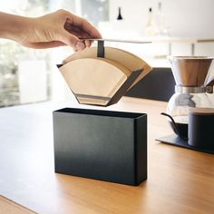 a person holding a coffee filter over a box on a wooden table next to a cup