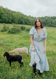 a woman standing in a field next to two sheep and one is looking at the camera