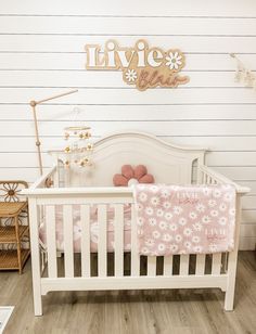 a white crib with pink bedding and flowers on the bottom, next to a wooden name sign