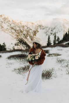 a woman in a white dress and fur coat standing in the snow holding a bouquet