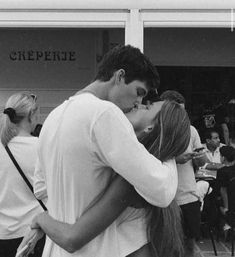 a man and woman kissing each other in front of a building with people standing around